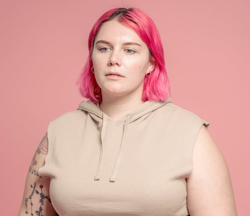 Emotionless young plus size female model wearing casual clothes standing against pink background and looking away